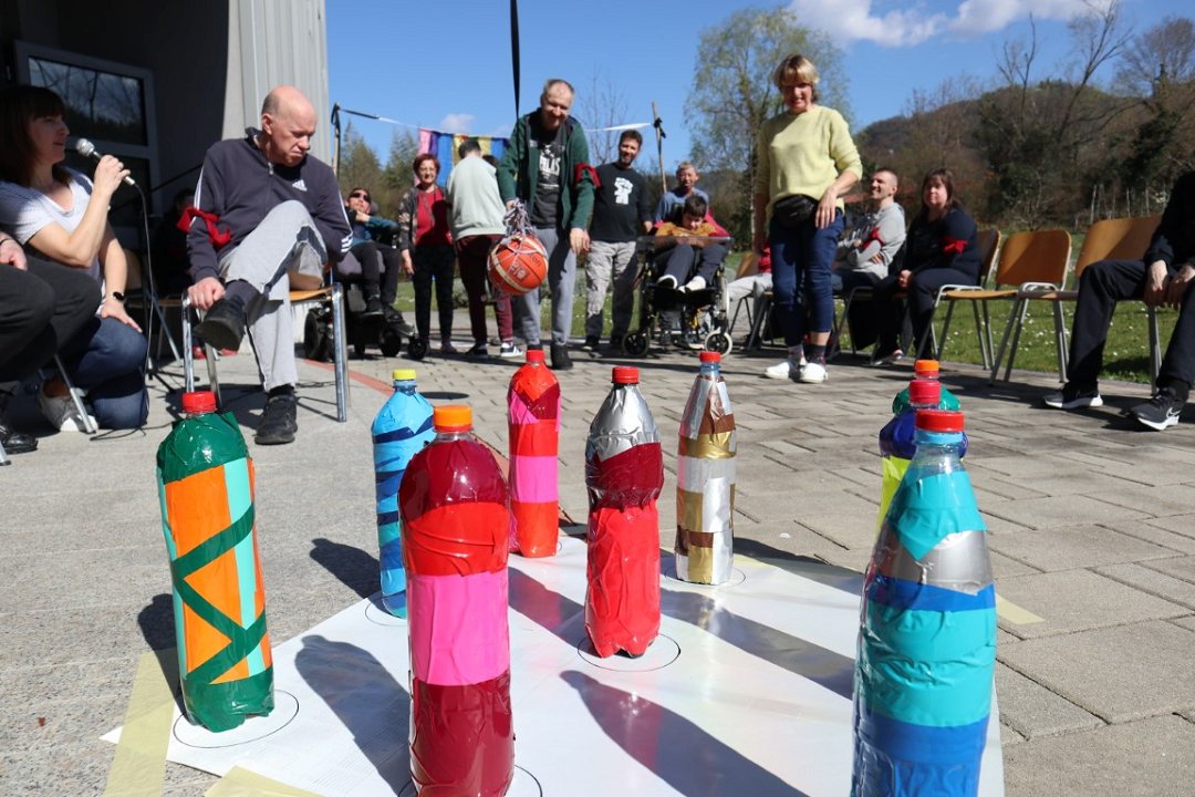 V ospredju fotografije je devet kegljev iz plastičnih steklenic, oblepljenih s pisanim papirjem. V ozadju je skupina uporabnikov in zaposlenih, Uporabnik drži v roki žogo in se pripravlja na met.