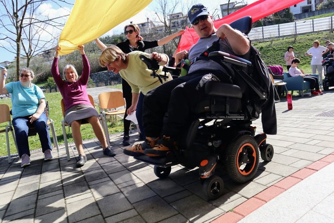 Uporabnik na električnem invalidskem vozičku v spremstvu zaposlene vozi pod pisanimi trakovi, ki jih držijo uporabniki in zaposleni.