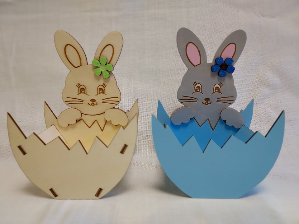 Na sliki sta dve košarici za čokoladna jajčka izdelani iz lesa. Zajček izrezan iz lesa drži med sprednjima tačkama spodnjo polovico jajčka, ki služi kot košarica. Levi zajček s košarico je v naravni barvi lesa in ima ob ušesu zeleno rožico, desni zajček s košarico je pobarvan v modro in ima ob ušesu modro rožico.